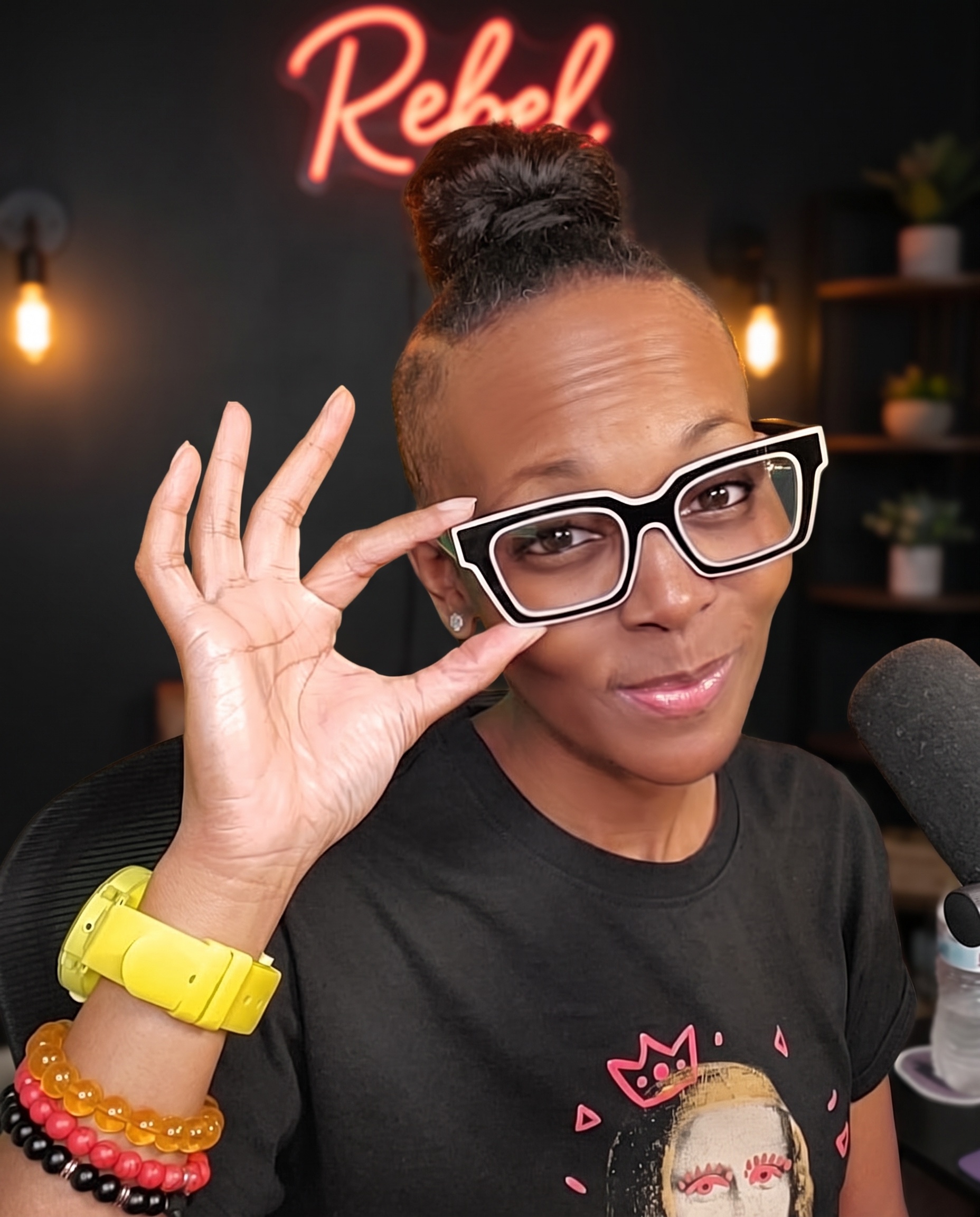 Rebel holding her glasses in her studio, red neon Rebel sign glowing behind her
